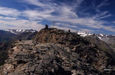 Das Bild zeigt den letzten und sehr steinigen Gratabschnitt zum Hohen Nebelkogel. Das große Gipfelkreuz ist bereits in Sicht. Bis dorthin führt der Steig zwischen den Gneisblöcken hindurch, wobei man ab und zu die Hände zur Unterstützung braucht. Am Gipfel stehen noch zwei große Steinmänner. Im Hintergrund sind die Gletschergipfel der Ötztaler Alpen zu erkennen. Direkt rechts neben dem Gipfel erhebt sich die Wildspitze. Es ist ein wunderschöner Sommertag, der Himmel ist blau und mit vielen Schleierwolken verziert.