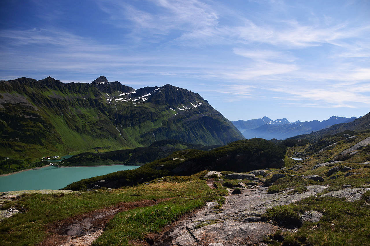 Rückblick von der Kammhöhe auf den Kops-Stausee, der milchig-türkis schimmert und das Zeinisjoch. Darüber erheben sich Fluhspitzen, Schrottenkopf und Fädnerspitze. Im Hintergrund zwei Dreitausender der Samnaungruppe: Vesulspitze und Bürkelkopf. Der blaue Julihimmel wird von Schleierwolken verziert. Im Vordergrund liegen große Gneisplatten neben dem Bergweg.