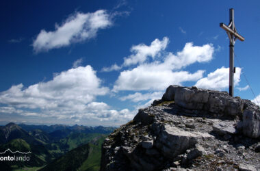 Am Gipfel der Namloser Wetterspitze nimmt der oberste Felskopf mit dem Kreuz die rechte Bildhälfte ein. Links im Hintergrund sind der Thaneller und die Ammergauer Alpen zu sehen - mehr grün als felsig. An diesem klaren Tag im Juli verzieren einige Schönwetterwolken den blauen Himmel.