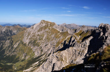 Auf dem Bild sieht man vom Nebelhorn hinüber zum Großen Daumen, dem höchsten Berg der gleichnamigen Gruppe. Zwischen den beiden Bergen erkennt man in der rechten Bildhälfte die Wengenköpfe, über die der Hindelanger Klettersteig verläuft. In der linken Bildhälfte ist die Rotspitze zu sehen und dahinter noch einige niedrige Voralpengipfel im Allgäu. Unter dem Großen Daumen liegt das Retterschwangtal. Die Berge hier sind felsig, zum Teil aber auch mit Gras bewachsen. An der braunen Färbung erkennt man, dass der Herbst nicht mehr weit ist. Der Himmel ist blau und wird nur von ganz wenigen Schleierwolken verziert. Die Fernsicht scheint an diesem klaren Tag fast unendlich.