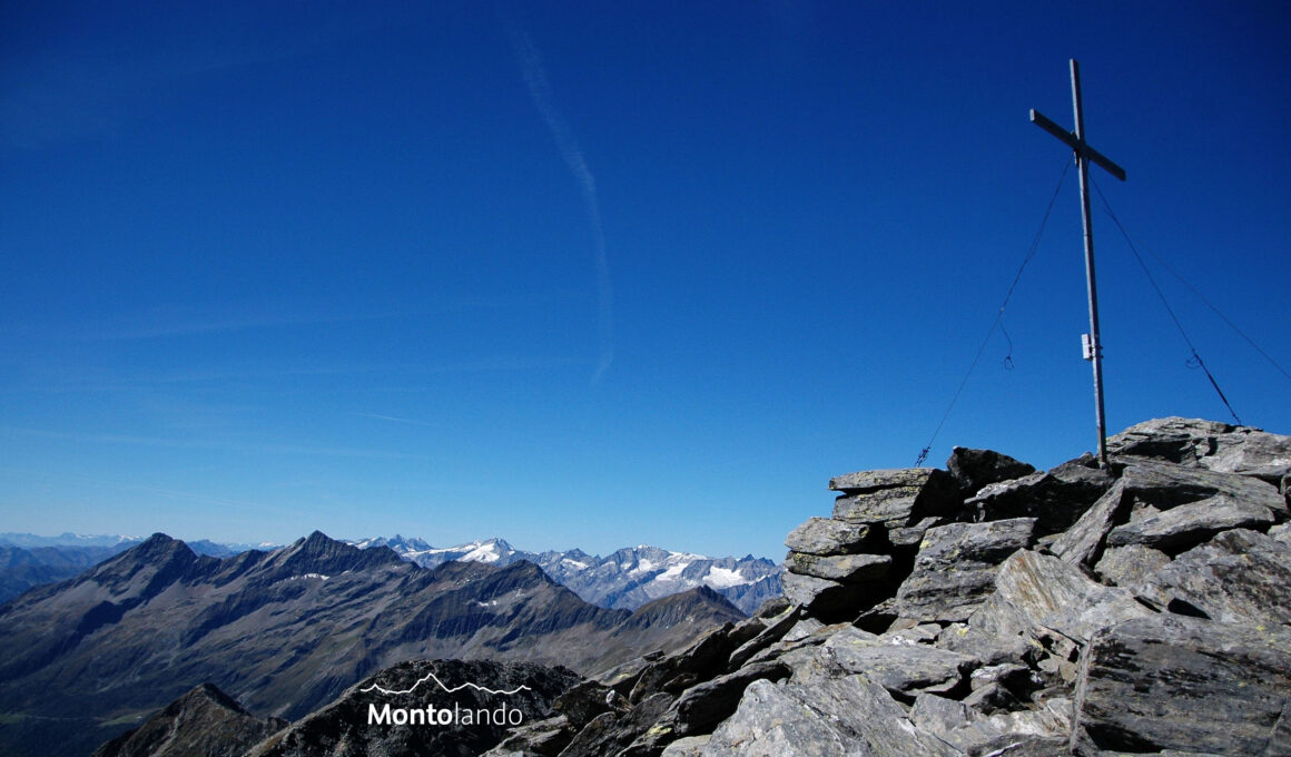 Auf dem Bild sieht man rechts auf dem Gipfelkopf der Dreieckspitze das große Gipfelkreuz. Es ist ein Traumtag und die Fernsicht scheint endlos zu sein. Das Gipfelkreuz steht auf einer Anhäufung von Gneisblöcken. In der linken Bildhälfte sieht man die Durreckgruppe und rechts dahinter den Hauptkamm der Zillertaler Alpen. Ganz links erkennt man in der Ferne die Ortlergruppe und etwas weiter recht die Wildspitze in den Ötztaler Alpen. Der stahlblaue Himmel nimmt geschätzt zwei Drittel des Bildes ein. Es hat nur ganz wenige Schleierwolken.