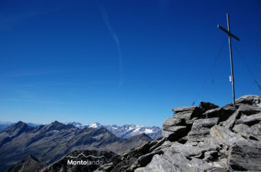 Auf dem Bild sieht man rechts auf dem Gipfelkopf der Dreieckspitze das große Gipfelkreuz. Es ist ein Traumtag und die Fernsicht scheint endlos zu sein. Das Gipfelkreuz steht auf einer Anhäufung von Gneisblöcken. In der linken Bildhälfte sieht man die Durreckgruppe und rechts dahinter den Hauptkamm der Zillertaler Alpen. Ganz links erkennt man in der Ferne die Ortlergruppe und etwas weiter recht die Wildspitze in den Ötztaler Alpen. Der stahlblaue Himmel nimmt geschätzt zwei Drittel des Bildes ein. Es hat nur ganz wenige Schleierwolken.