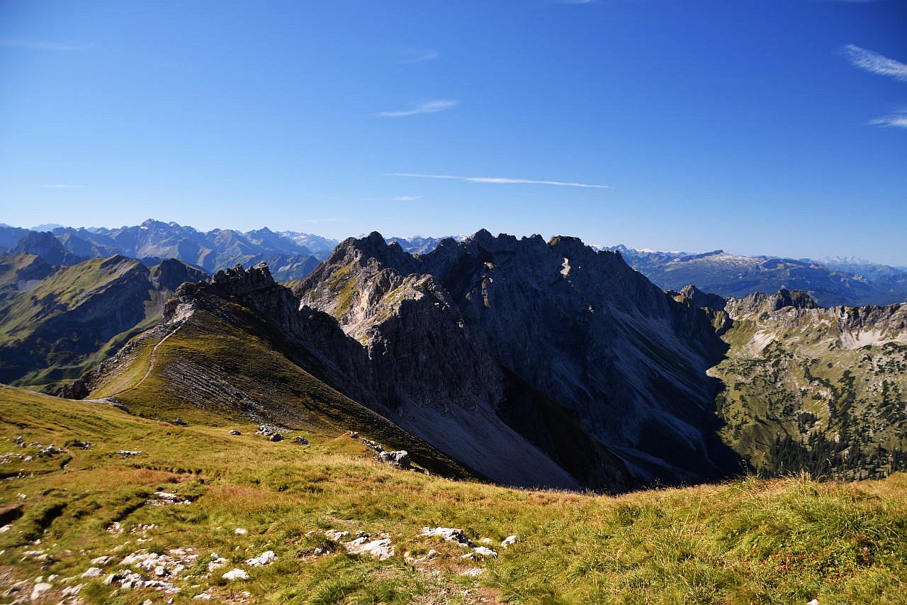 Vom Schlussanstieg zum Großen Daumen sieht man hinüber zum Grat zwischen Wengenköpfen und Nebelhorn. Die schattigen Nordwände auf der rechten Seite zeigen das Gestein, den Hauptdolomit. Darunter liegen große Schutthalden. Im Hintergrund erheben sich am Horizont viele Gipfel der Allgäuer Alpen von den Höfats ganz links, über Mädelegabel, Biberkopf zum Hohen Ifen rechts. Am rechten Bildrand erkennt man in der Ferne noch das Säntismassiv in der Schweiz. Im Vordergrund erstreckt sich die Hochweide am Glasfeld. Der Himmel ist blau und wird nur von ganz wenigen Schleierwolken verziert.