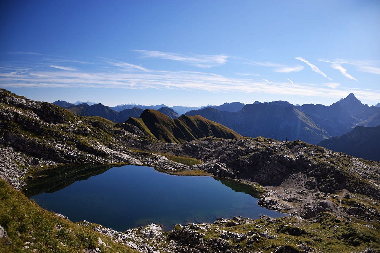 Auf dem Bild sieht man von etwas oberhalb, vom Steig zum Großen Daumen, auf den Laufbichelsee hinab. Der See liegt in einer Mulde und auf einer kleinen Anhöhe südlich (im Bild rechts vom See), steht ein Kreuz. Im Hintergrund erheben sich viele Gipfel der Allgäuer Alpen. Ganz rechts erkennt man den markanten und formschönen Hochvogel. Der Himmel ist blau und wird nur von ganz wenigen Schleierwolken verziert.