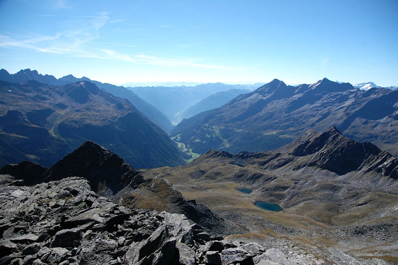 Vom Bärenluegspitz sieht man hinab in die Mulde, in der die Kofler Seen liegen. Diese wird von spitzigen Gneisgipfeln eingerahmt. In der Verlängerung der Mulde liegen in der Tiefe das Reintal und weiter hinten das Mühlwalder Tal. Beide Täler werden ebenfalls von mächtigen Gneisgipfeln eingerahmt. Am Horizont verblauen die Berge im Gegenlicht der Nachmittagssonne. Der Himmel ist blau und wird von wenigen Schleierwolken verziert.