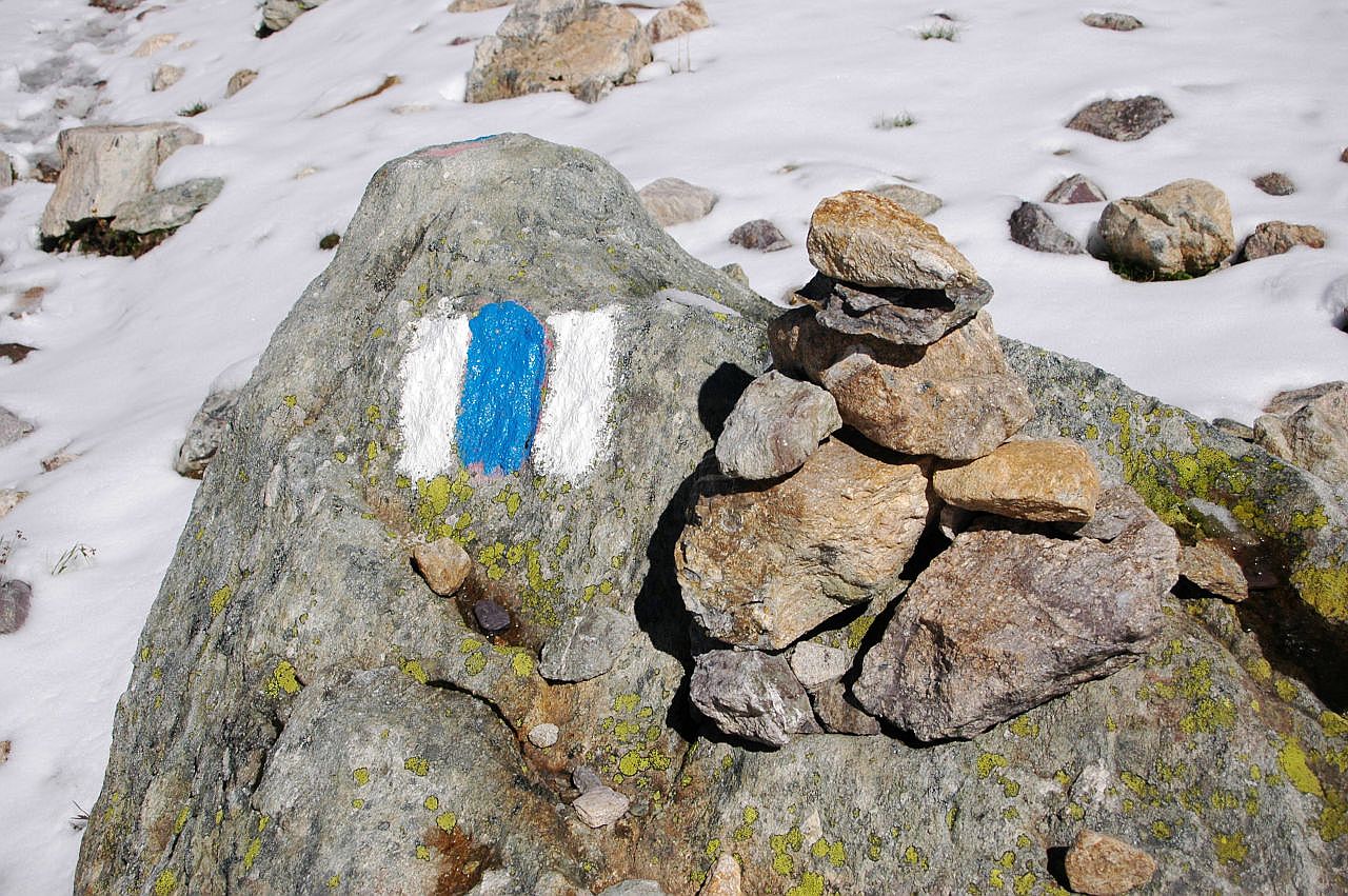 Auf dem Bild sieht man eine weiß-blau-weiße Markierung, die auf einen Felsen gemalt ist. In der Schweiz werden so genannte Alpinwanderrouten auf diese Weise gekennzeichnet. Rechst neben der Markierung steht auf dem Felsen noch ein kleiner Steinmann. Das Gelände im Hintergrund ist schneebedeckt, es ragen nur wenige Felsen heraus.