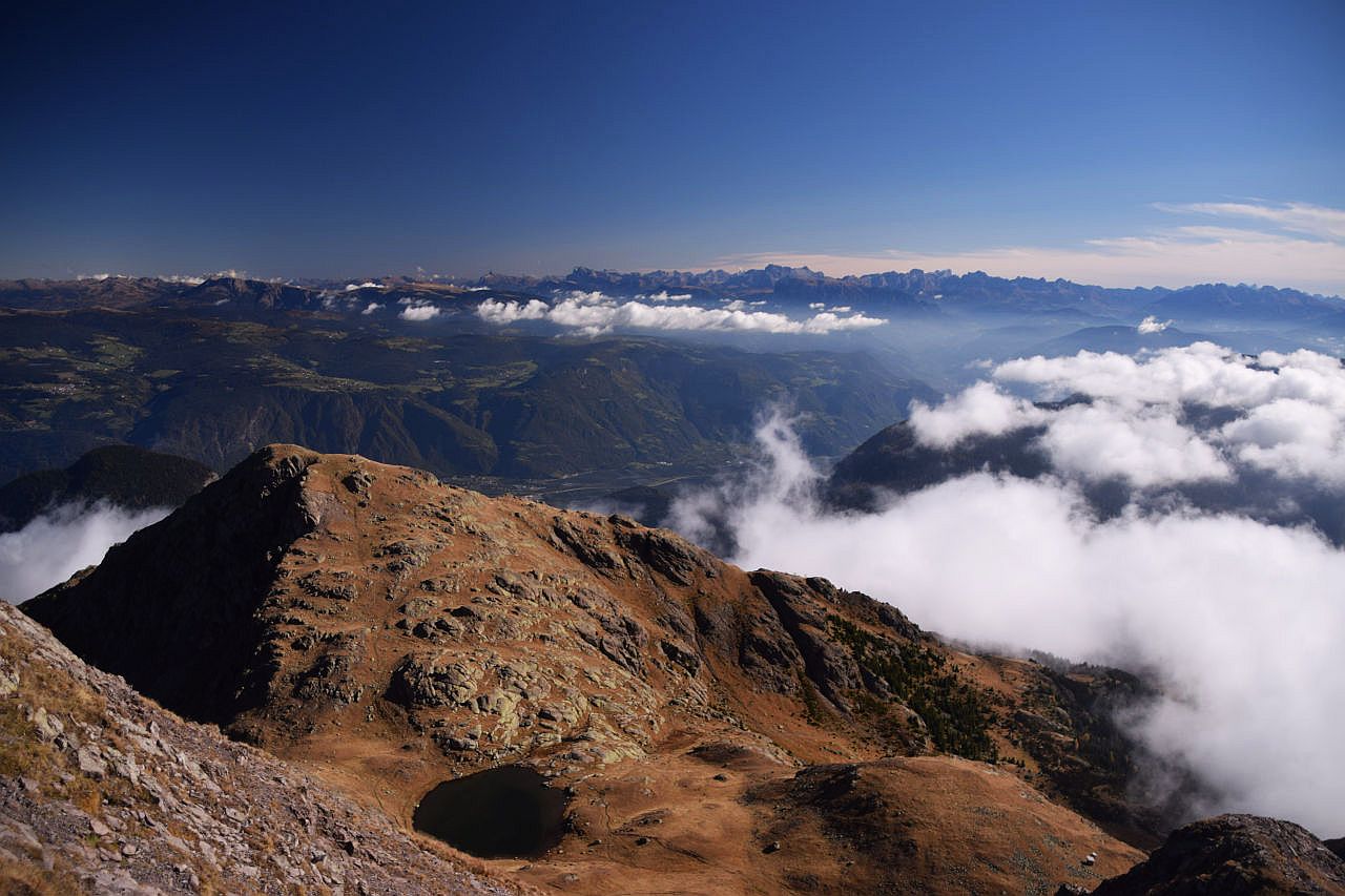 Auf dem Bild geht der Blick über den Laugensee, der ca. 250 Meter tiefer liegt und die Kleine Laugenspitze hinweg zu den Bergen der Sarntaler Alpen und den Dolomiten, die sich am Horizont erheben. Rechts sind noch einige Berge des Lagorai erkennbar. Zwischen den Gipfel ziehen Wolkenschwaden entlang. Vor allem rechts von der Kleinen Laugenspitze verdeckt eine größere Wolke den Blick auf den Gampenpass und den Mendelkamm. In der rechten Bildhälfte hat es am Horizont Schleierwolken. Ansonsten ist der Himmel blau und die Fernsicht scheint grenzenlos.