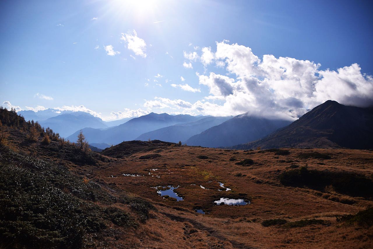 Auf dem Bild blickt man vom Anstieg zur Laugenspitze oberhalb der Aler Alm Richtung Süden über das Nonstal hinweg Richtung Val di Sole im Trentino. Im Vordergrund sieht man ein Hochmoor, das von ein paar Pfützen geschmückt wird und bereits herbstlich gefärbt ist. Im Hintergrund sieht man links die Gipfel der Brentagruppe, in der Bildmitte und rechst die Magdalener Berge. Dort und an der Brentagruppe hängen einige Wolken, die manche Gipfel einhüllen. Ansonsten ist der Himmel blau und sogar die Sonne ist am oberen Bildrand noch teilweise zu sehen.