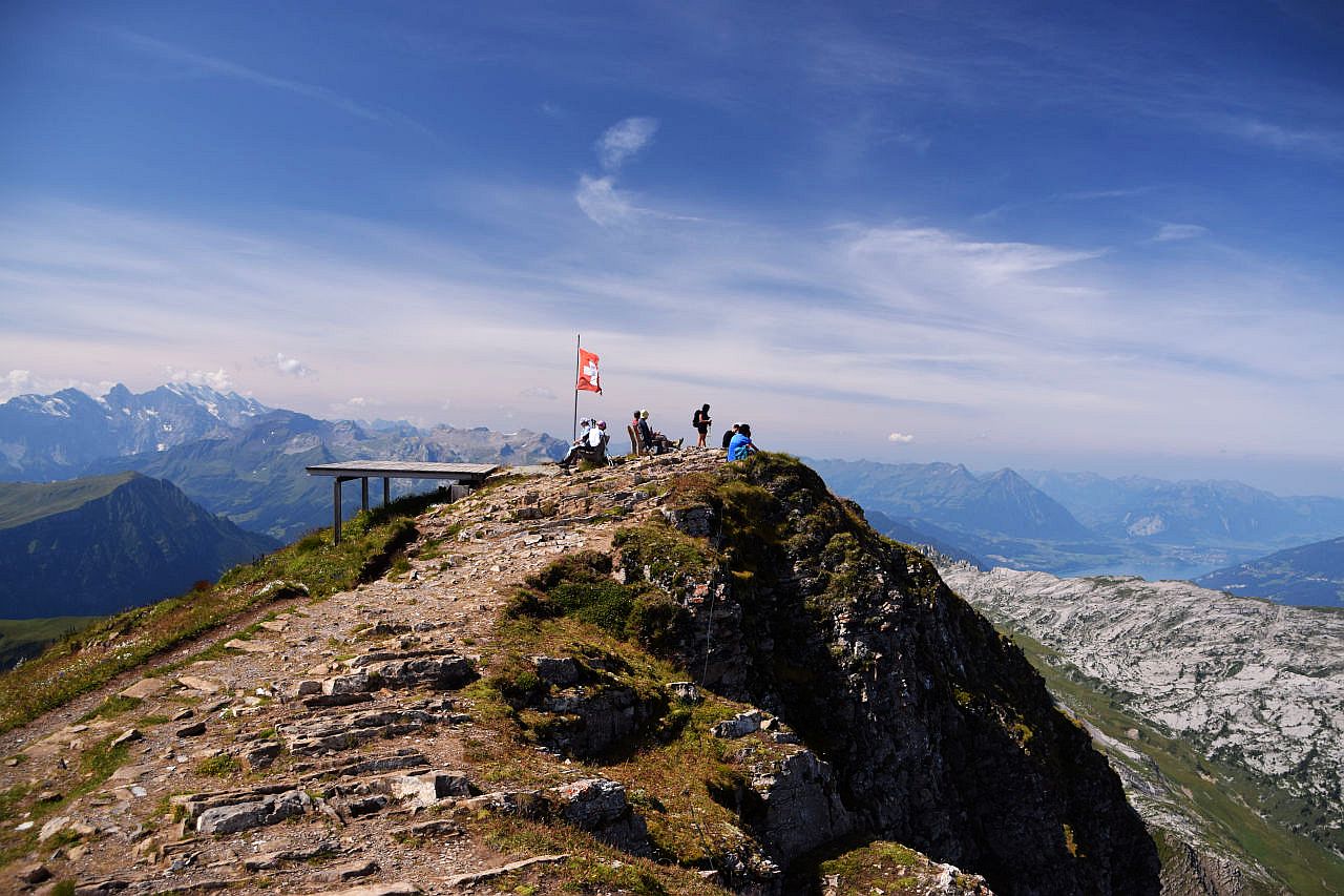 Auf dem Bild geht der Blick über den Gipfel des Faulhorns Richtung Westen. Auf dem Gipfel sitzen mehrere Bergwanderinnen und Bergwanderer auf Ruhebänken oder auf Steinen. Dahinter ist eine Fahne mit dem Schweizer Kreuz zu sehen und links einen Startplatz für Drachensegler. Im Hintergrund sieht man links die Berner Hochalpen mit dem Blüemlisalpmassiv, rechts sieht man die Berner Voralpen mit dem Niesen und dem Thunersee. Der Himmel ist blau, aber zum Teil von dichten Schleierwolken bedeckt.