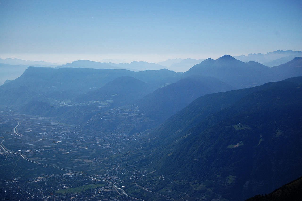 Auf dem Bild sieht man von den Höhen der Texelgruppe oberhalb von Meran zu den beiden Laugenspitzen in der rechten Bildhälfte und zum Mendelkamm links. Zwischen beiden Massiven erkennt man d den Gampenpass. Hinter den Laugenspitzen sieht man am Horizont noch die Brentagruppe und am linken Bildrand wenige Gipfel der Fleimstaler Alpen. Es handelt sich um eine Gegenlichtaufnahme, die Felsstrukturen der Berge sind daher kaum sichtbar. Die Gipfel verblauen in der Ferne. In der Tiefe sieht man im Vorder- und Mittelgrund das Etschtal bei Meran mit Lana und Marling. Der Himmel ist blau, aber zwischen den Bergen steigt Dunst empor, was auf einen Wetterwechsel hindeuten könnte.