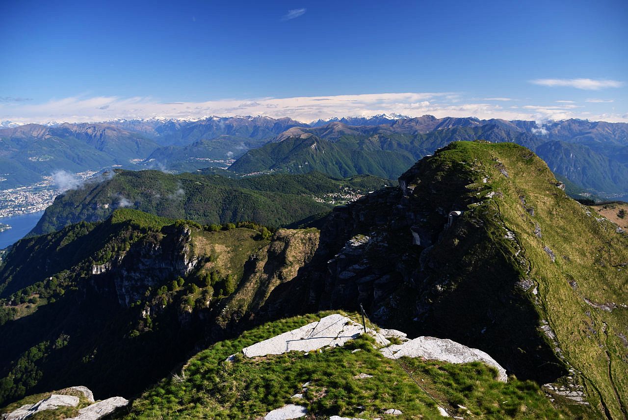 Auf dem Bild blickt man über den nördlichen Vorgipfel des Monte Generoso, den Baraghetto, hinweg nach Norden. Es ist ein klarer Tag mit Nordföhn und es sind sehr viele Gipfel erkennbar. Die niedrigeren Gipfel im Vordergrund sind im Mai bereits grün, während die hohen Gipfel der Adula und der Tessiner Alpen im Hintergrund zum Teil noch schneebedeckt sind. Links am Bildrand ist noch in Teil von Lugano und ein kleiner Ausschnitt des Luganersees erkennbar. Der Himmel ist blau und wolkenlos. Nur am Horizont hat es dichte Wolken. Diese deuten an, dass das Wetter auf der Alpennordseite nicht so schön ist.