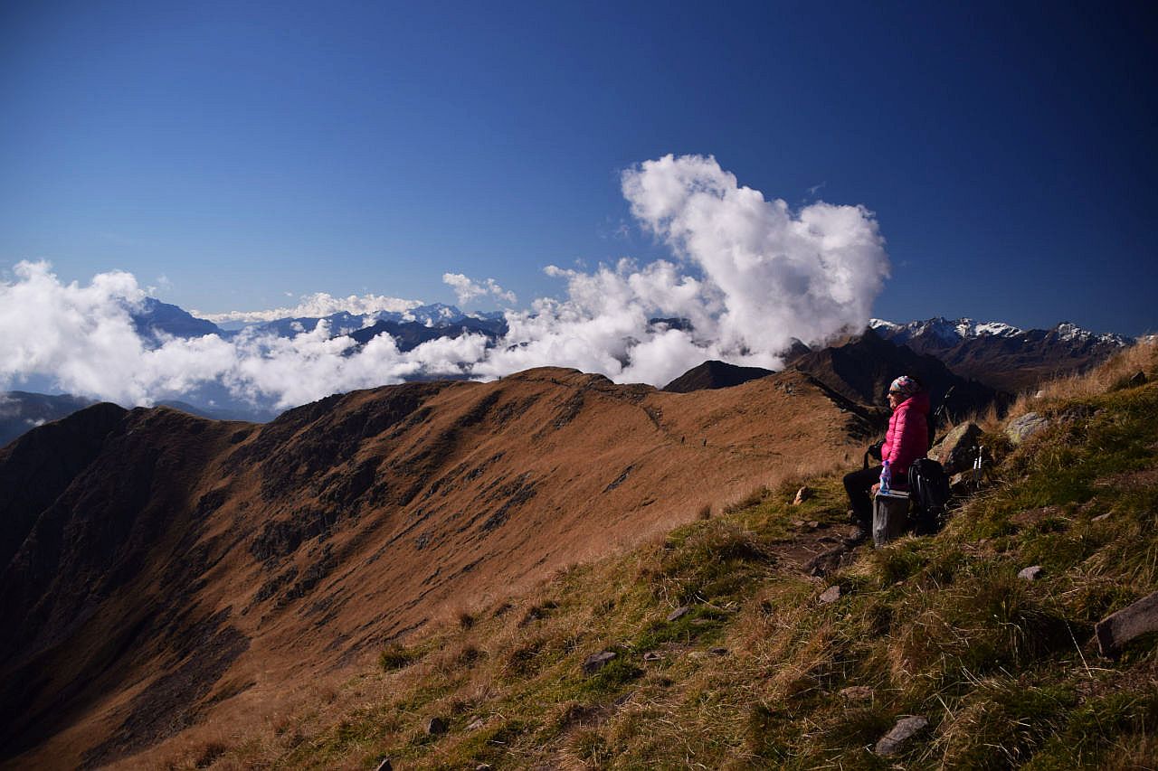 Auf dem Bild sieht man eine Bergwanderin, die am Gipfel der Laugenspitze auf einer Bank sitzt und Richtung Süden in die Sonne schaut. Im Mittelgrund sieht man den langen Grat, der vom Gipfel nach Süden zieht mit der Wegspur und mehreren Bergwanderern. Dahinter hat es Quellwolken, von denen die Berge im Hintergrund verdeckt werden. In der Bildmitte schaut gerade noch die Cima Presanella heraus, rechts erkennt man über der Bergwanderin noch einen Teil des Zufrittkamms. Über den Quellwolken ist der Himmel blau.