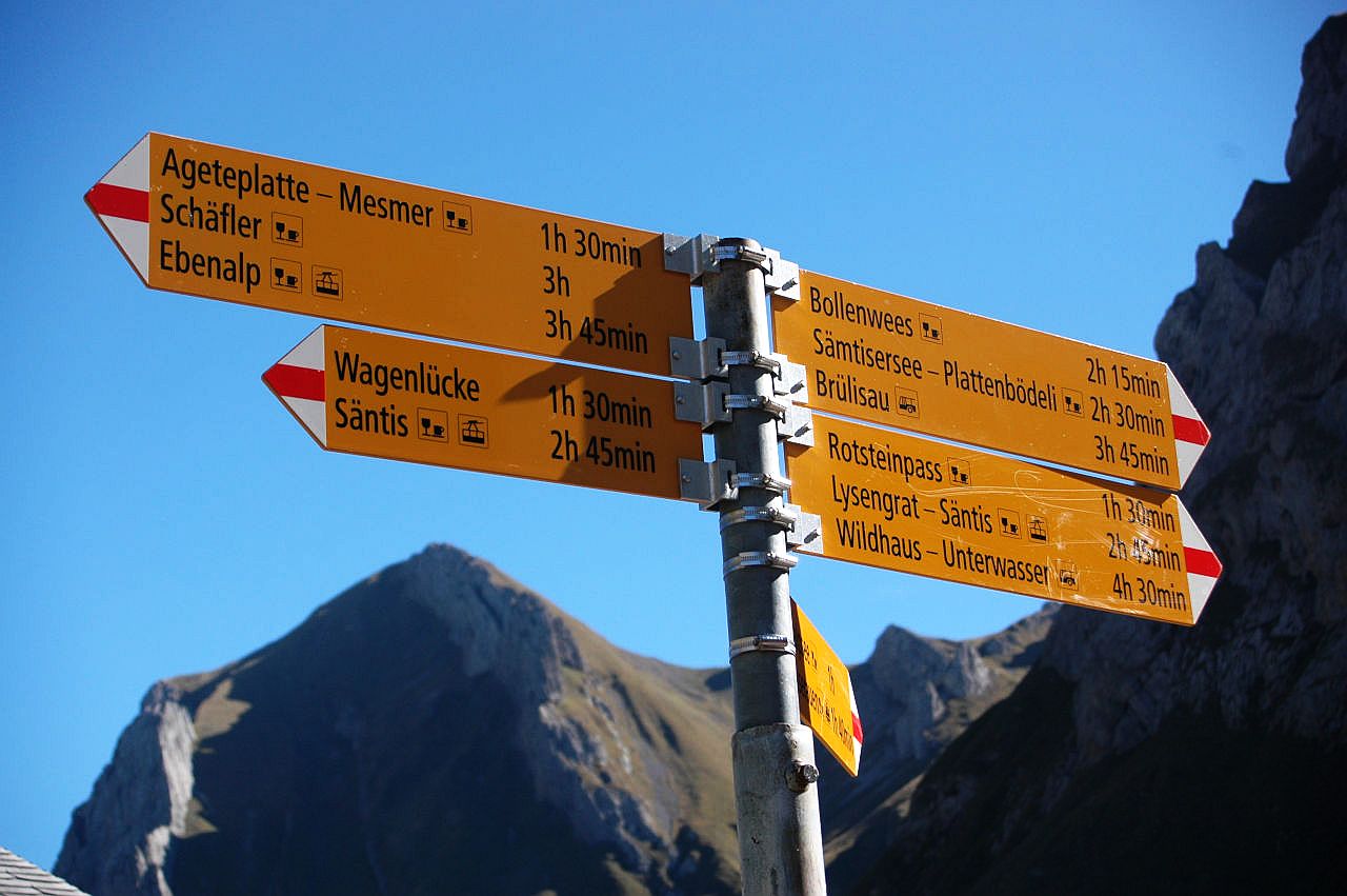 Das Bild zeigt einen gelben Wegweiser für Bergwanderwege in der Schweiz. Nach links und rechts zeigen Richtungspfeile verschiedene Wanderziele und die Wanderzeiten an. Die Pfeilspitzen sind weiß-rot-weiß, den Farben für Bergwanderwege. Der Himmel ist blau wolkenlos. Die Berge im Hintergrund sind nur verschwommen erkennbar.