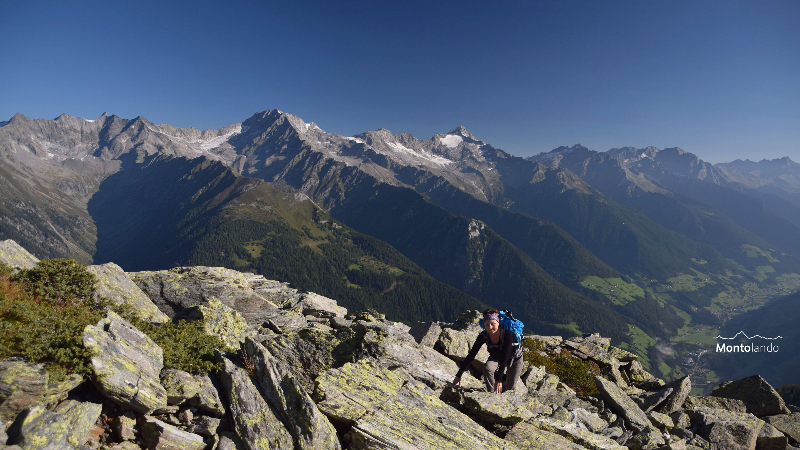 Das Bild zeigt eine Wanderin beim Anstieg durch ein Blockfeld aus großen Gneisblöcken. Im Hintergrund erheben sich die Gipfel des Zillertaler Hauptkammes über dem Ahrntal, das auf der rechten Seite zu sehen ist. Am einem Traumtag Ende August ist keine Wolke am Himmel zu sehen.