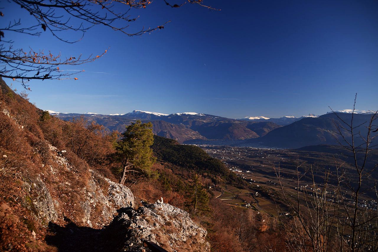 Auf dem Bild geht der Blick vom Alten Mendelweg Richtung Norden. Im Mittelgrund erkennt man rechts die Streusiedlung Eppan und dahinter, noch im Schatten, Teile von Bozen. Darüber erheben sich im Hintergrund die flachen Berge der Sarntaler Alpen. Sie sind nur in den Gipfelregionen schneebedeckt. Ganz rechts schauen noch ein paar Gipfel der Dolomiten heraus. Im Vordergrund links zieht der mit Sträuchern und Bäumen bewachsene Hang zum Mendelkamm hinauf. Im Vordergrund liegen einige Kalkfelsen, zwischen denen der Weg verläuft. Der Himmel ist blau, klar und fast wolkenlos.