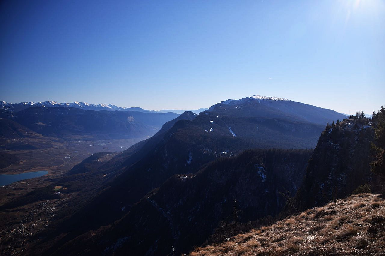 Auf dem Bild blickt man von der im Text erwähnten Hochfläche auf der Südseite des Penegal Richtung Süden. Links in der Tiefe liegen Etschtal und Kalterer See. Darüber baut sich im Hintergrund das Trudener Horn auf, darüber der schneebedeckte Lagorai. In der rechten Bildhälfte sieht man die bewaldeten Nordflanken des Mendelkamms, aus dem als höchster Punkt der im Gipfelbereich schneebedeckte Monte Roen herausragt. Rechts am Bildrand erkennt man im Gegenlicht das im Text erwähnte Kreuz. Der Himmel ist blau und wolkenlos.