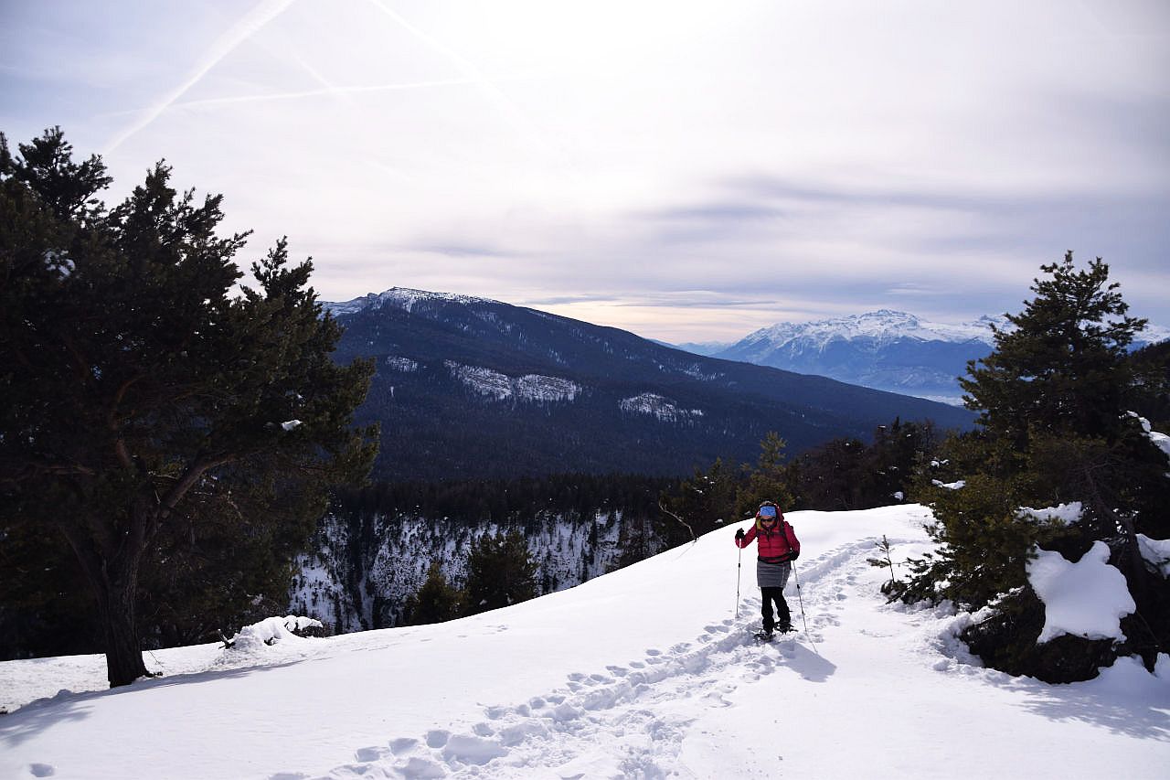 Auf dem Bild sieht man eine Wanderin mit Schneeschuhen, die in wenigen Augenblicken das im Text erwähnte Kreuz erreichen wird. Der Anstieg ist gespurt, es waren vor ihr wohl schon andere Schneeschuhwanderer hier. Links und rechts von der Spur hat es Kiefern. Im Hintergrund erkennt man links den Monte Roen und rechts die Brentagruppe. Der Himmel ist von dichten Schleierwolken bedeckt, die Sonne schient nur milchig hindurch.