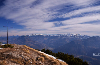 Auf dem Bild geht der Blick vom Nordgipfel der Marzola in nordöstlicher Richtung zu den Bergen des Lagorai. Man sieht die Gipfel rund um die Val dei Mocheni (Fersental), vom Dosso di Costalta über Monte Ruioch, Monte Gronlait, Monte Fravort zur Cima Panarotta am rechten Bildrand. Am linken Bildrand ragt das Gipfelkreuz in den blauen Himmel. Die Berge sind nur in den Gipfelregionen schneebedeckt. Links sind hinter dem Kreuz erkennt man am Horizont noch einige Gipfel in Südtirol, die deutlich mehr Schnee tragen. Der blaue Himmel ist mit dichten Schleierwolken bedeckt. Die Sonne scheint aber hindurch und trotz der Wolken ist der Gesamteindruck freundlich.