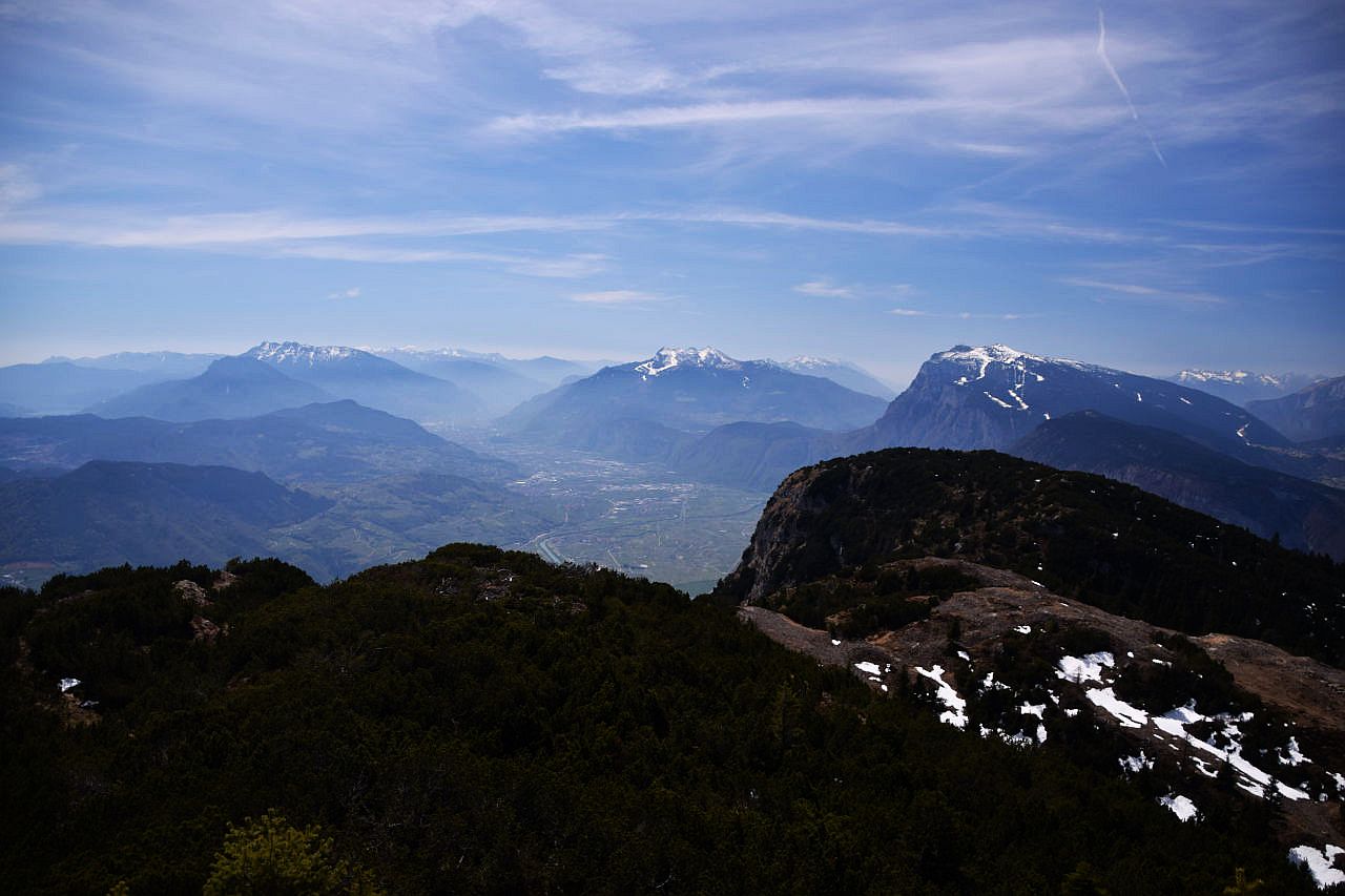Auf dem Bild geht der Blick von der Cima Monticello, einem Berg im südlichen Mendelkamm über einen Vorgipfel hinweg Richtung Süden zu den Bergen des Trentino. Genau in der Bildmitte erkennt man das dicht besiedelte Etschtal zwischen Mezzocorona und Trento. Links darüber die Vizentiner Alpen mit der Marzola und der Vigolana. Weiter rechts anschließend die Gipfel des Monte Bondone, die Paganella und ganz rechts die Gardaseeberge. Fast alle Berge sind in den Gipfelregionen noch schneebedeckt. Der Himmel ist blau und mit Schleierwolken verziert.