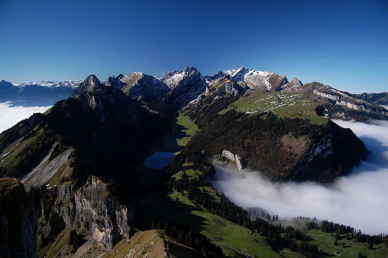 Auf dem Bild blickt man vom Hohen Kasten auf den Alpstein. Am Horizont erkennt man die beiden höchsten Gipfel, Altmann und Säntis mit Antenne. In der Bildmitte erkennt man im Schatten den Sämtisersee. In der rechten Bildhälfte breitet sich unterhalb des Säntis die Hochfläche der Alp Sigel aus. Von rechts ziehen Nebelschwaden aus nördlicher Richtung in die Täler des Alpsteins. Der Himmel ist blau und wolkenlos. Es ist ein außergewöhnlich klarer Herbsttag.