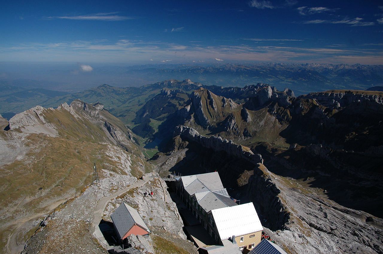 Auf dem Bild geht der Blick vom Säntis über das Berggasthaus und den Alpstein hinweg zu den Bergen in Vorarlberg. Ziemlich genau in der Bildmitte erkennt man in der Tiefe den Seealpsee, über dem links oberhalb Ebenalp und Schäfler zu sehen sind. In der hintersten Bergkette des Alpsteins sieht man auch den Hohen Kasten. Dahinter erkennt man das Rheintal, über dem ein leichter Dunstschleier liegt. Darüber sieht man am Horizont unzählige Gipfel in Vorarlberg. Der Himmel ist blau und wird von schönen Schleierwolken verziert.