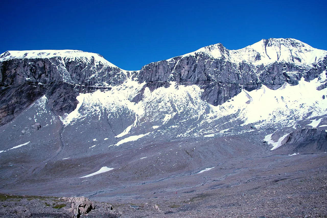 Auf dem Bild sieht man die Glarner Hauptüberschiebung am Piz Segnas von Osten, vom Weg zum Trinserhorn. Wie ein Strich zieht sich die Grenze der Überschiebung durch die gesamte Ostflanke des Piz Segnas, der am rechten Bildrand zu sehen ist. Nachlinks zieht der Grat zum Atlas. Beide Gipfel sind mit Neuschnee bedeckt. Im Vordergrind sieht man die Schuttmulde, die man im Anstieg zum Sardonapass und Trinserhorn durchquert. Der Himmel ist blau und wolkenlos.