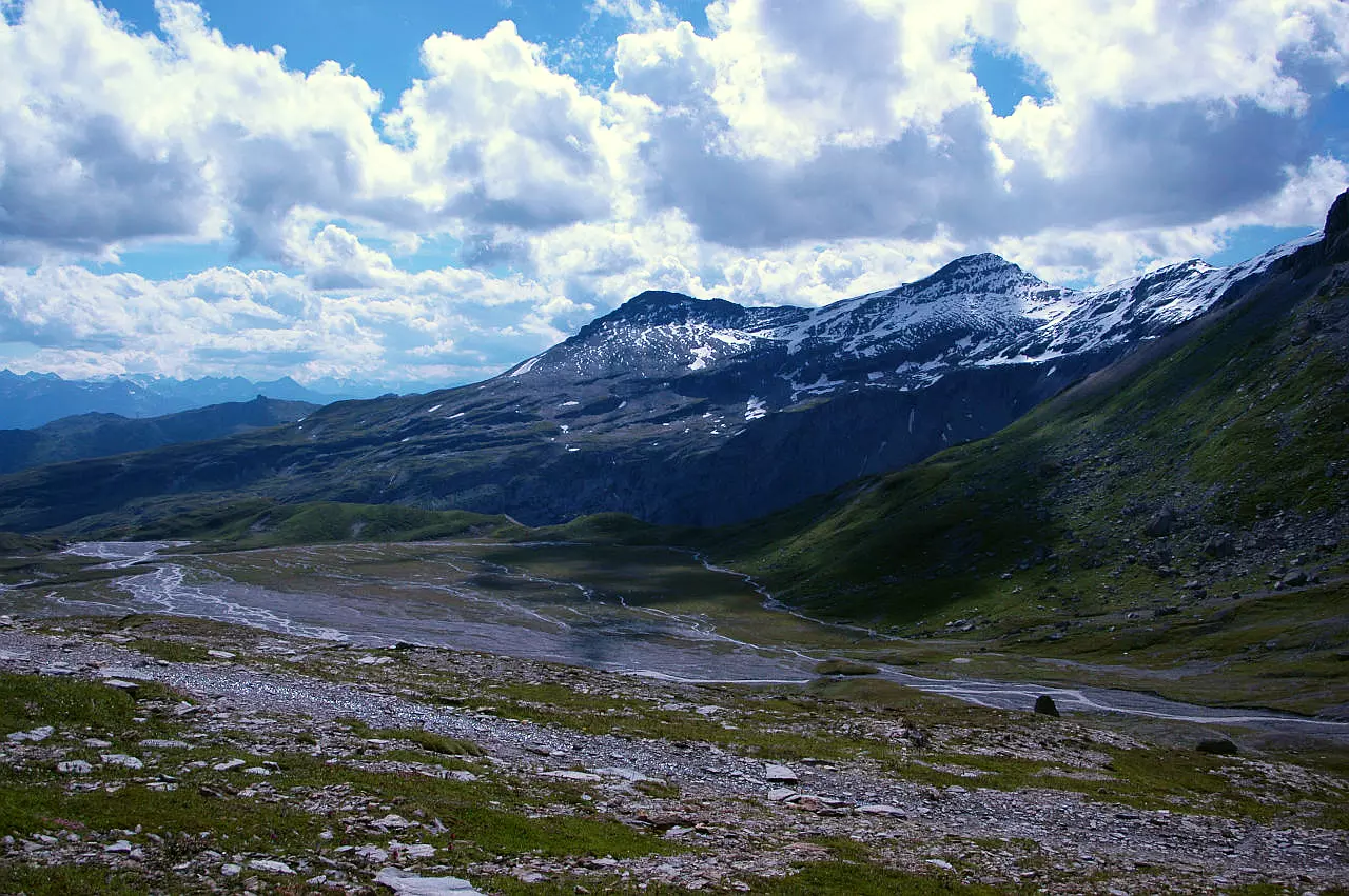 Auf dem Bild geht der Blick vom Anstieg zum Trinserhorn hinab auf die Hochebene Plaun Segnas Sura, den Oberen Segnasboden. Die Hochebene wird von vielen kleineren Bächen durchquert, die sich aber im Hintergrund zu einem größeren Bach vereinigen. Im Hintergrund sieht man rechts das Laaxer Stöckli und seinen Vorgipfel, den Crap Ner. Links am Horizont erkennt man die Gipfel der Adula. Der Himmel ist blau, aber es hat viele Quellwolken. Sie decken die Sonne zum Teil ab und werfen Schatten auf die Landschaft.