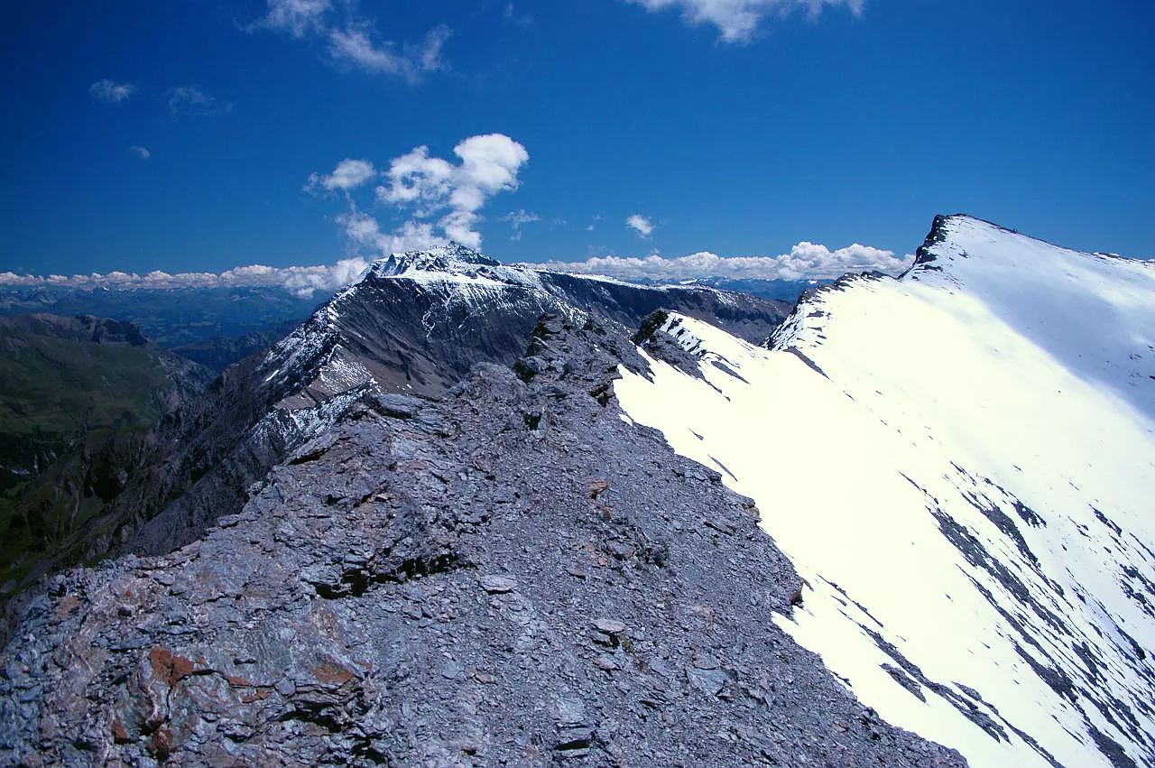 Auf dem Bild geht der Blick vom Nordwestgipfel des Trinserhorns zum Hauptgipfel, der man rechts hinten sieht. Im Vordergrundsieht man links den brüchigen Grat, der zum Hauptgipfel zieht. Die nach rechts (Süden) abfallende Flanke ist mit Neuschnee bedeckt und der Steig, der zum Hauptgipfel führt ist deshalb nicht sichtbar. Links erkennt man im Hintergrund über dem Grat den Ringelspitz und dahinter in er Ferne noch viele Gipfel der Bündner Alpen. Links am Bildrand sieht man noch einen Ausschnitt vom Calfeisental. Der Himmel ist blau, es hat wenige Quellwolken.