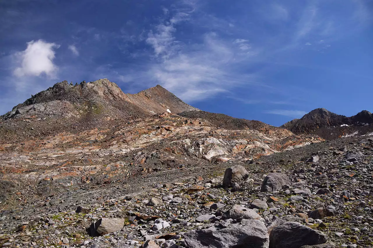 Auf dem Bild sieht man den Scheiblehnkogel von Nordwesten, aus dem großen geröllreichen Kar auf der Westseite des Gipfels, das der Anstieg durchquert. Der Gipfel erhebt sich im Hintergrund als flache Pyramide. Der im Text erwähnte Sattel, P. 2888m, wird von einem Buckel verdeckt. Die Landschaft besteht hier aus Blockhalden & Gletscherschliffen, also das, was der Gletscher zurückgelassen hat. Rechts im Bild erkennt man noch eine Moräne, die mit etwas Gras bewachsen ist. Ansonsten wirkt alles hier rau und herb. Der Himmel ist blau, es hat dünne Schleierwolken.