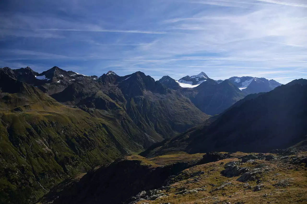 Auf dem Bild geht der Blick vom Weg zum Brunnenkogel über das Windachtal hinweg. Gegenüber erhebt sich eine eindrucksvolle Bergkette der Stubaier Alpen, von den Daunkögeln links, über die Stubaier Wildspitze und Schaufelspitze zum Zuckerhütl und zur Sonnklarspitze ganz rechts. Man erkennt noch ein paar kleinere Gletscher und die höchsten Gipfel haben en wenig Neuschnee. Der Talboden des Windachtals ist nicht sichtbar. Der Himmel ist blau, es hat aber viele dünne Schleierwolken.