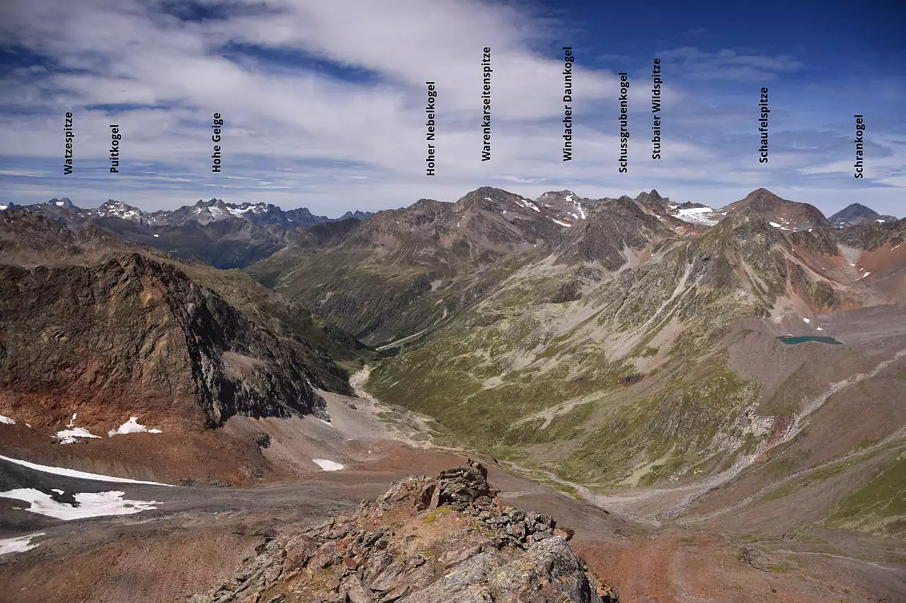 Auf dem Bild geht der Blick vom Scheiblehnkogel, einem leichten 3000er über das weitläufige Windachtal hinweg zu den Gipfeln nördlich des Tal zwischen Hohem Nebelkogel und Schaufelspitze. Vom Gipfel ist der Ausgangspunkt der Wanderung beim Gasthaus Fiegl nicht sichtbar. Links im Hintergrund schauen noch einige Gipfel der Ötztaler Alpen mit Watzespitze und Hoher Geige hervor, rechts am Bildrand der Schrankogel. Auffallend sind viele rötliche Gesteine, was auf einen hohen Eisengehalt schließen lässt. Sie geben der Landschaft ein buntes Aussehen. Der Himmel ist blau, es hat viele schöne Schleierwolken.