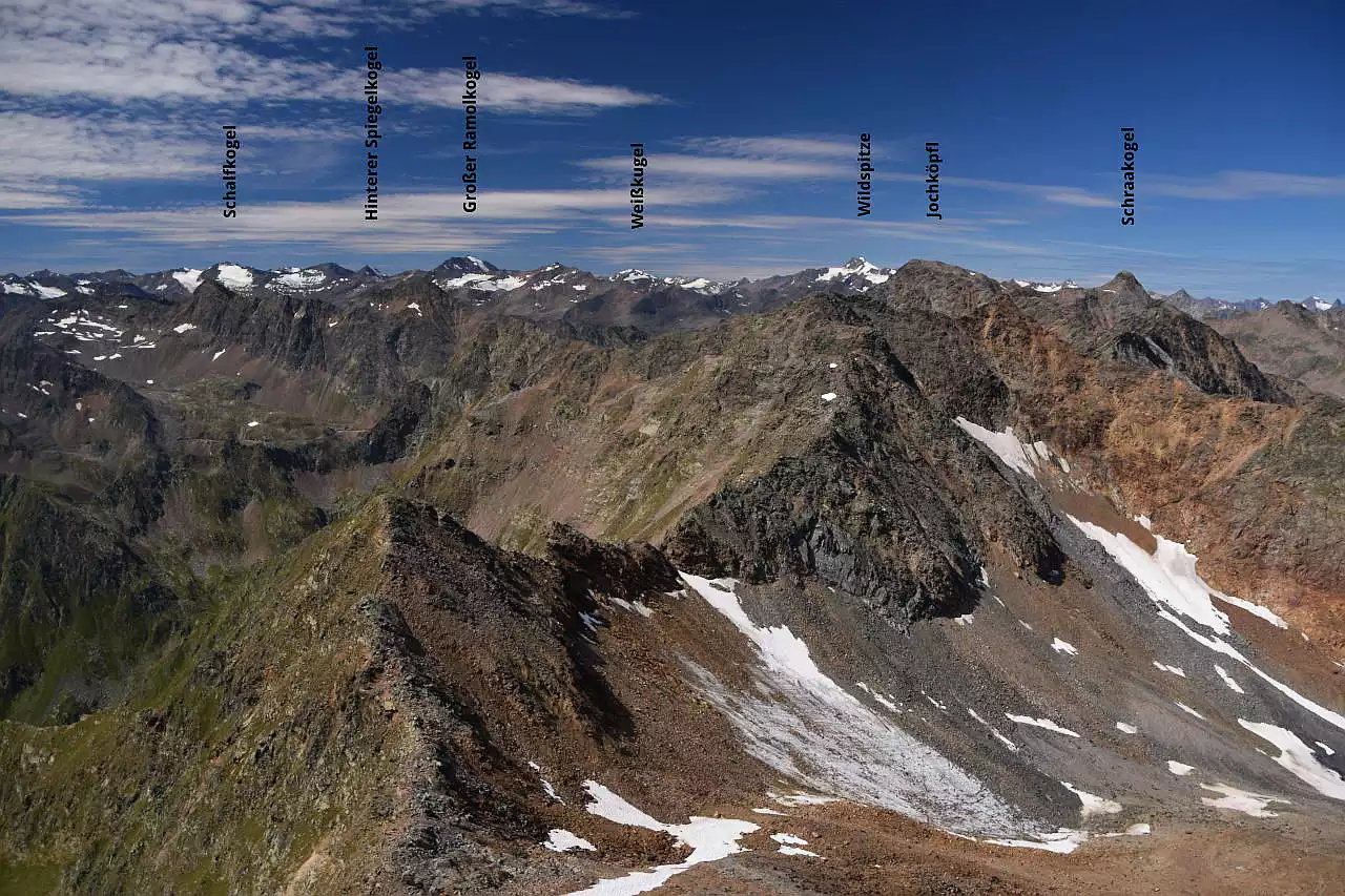 Auf dem Bild geht der Blick vom Scheiblehnkogel, einem leichten 3000er nach Westen zu den Gipfeln im Windachkamm im Vordergrund und den Ötztaler Alpen am Horizont. Rechts unten sieht man das blockgefüllte Kar, durch das der Anstieg zum Scheiblehnkogel erfolgt. Man sieht auch noch die Reste des Westlichen Scheiblehnferners, der in weiten Teilen aber mit Geröll bedeckt ist. Die Gesteine der Gipfel im Windachkamm sind zum Teil rötlich, auf der linken Seite fallen steile, teils begrünte Hänge Richtung Süden ins Timmelstal ab. Die Gipfel der Ötztaler Alpen sind teils vergletschert, vor allem die Wildspitze, der höchste Berg im Ötztal. Der Himmel ist blau und es hat schöne Schleierwolken.