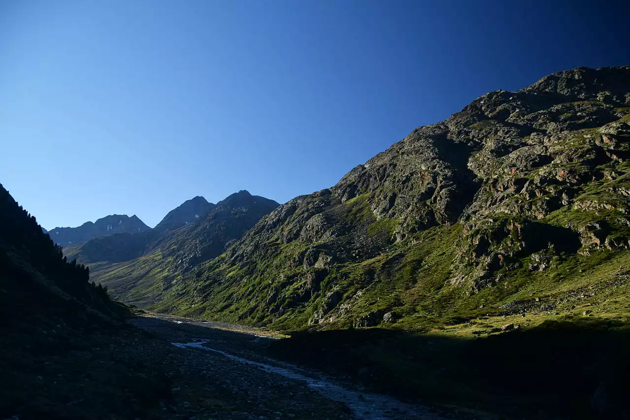 Auf dem Bild geht der Blick ins Windachtal taleinwärts. Über dem den Tal erheben sich im Hintergrund die Gipfel im Windachkamm. Ganz hinten links erkennt man die kleine Pyramide des Scheiblehnkogels, rechts neben der breiten Peillöcherspitze. Im Vordergrund erkennt man die Windache, die durch das gleichnamige Tal fließt. Sie liegt noch im Schatten, während auf die Gipfel bereits die Sonne fällt. Der Himmel ist blau und wolkenlos.