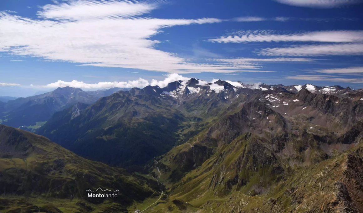 Auf dem Bild geht der Blick vom Scheiblehnkogel Richtung Süden und Südwesten. Links sieht man die Gipfel im Gurgler Kamm in den Ötztaler Alpen. Sie sind in der Gipfelregion vergletschert. Links sieht man die östlichen Ausläufer der Texelgruppe mit der Kolbenspitze und in der Ferne links dahinter noch die Berge im Meraner Land. m Vordergrund liegt in der Tiefe das Timmelstal. Der Himmel ist blau, es hat Schleierwolken, die den Himmel wunderschön verzieren.