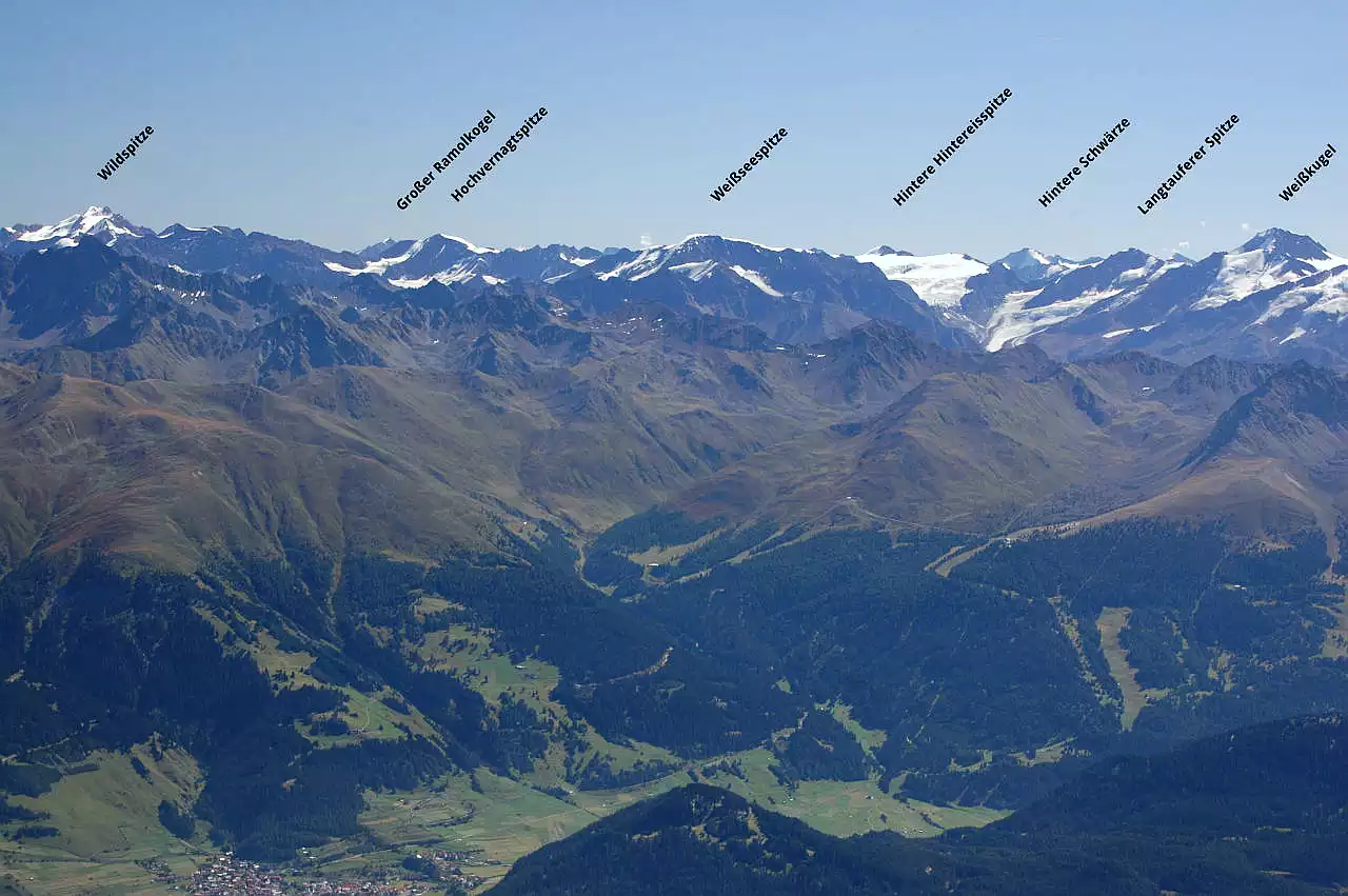 Auf dem Bild geht der Blick vom Muttler Richtung Südosten zu den 3000ern der Ötztaler Alpen. Ganz links die Wildspitze, der höchste Berg der Gruppe und ganz rechts die Weißkugel der zweithöchste Berg. Dazwischen sieht man noch viele bedeutende Gipfel wie die breite Weißseespitze in der Bildmitte. Unten links erkennt man noch im Vordergrund Nauders am Reschenpass und an den Wiesenhängen die Skilifte. Es ist ein außergewöhnlich klarer Tag. Der Himmel ist blau und wolkenlos.