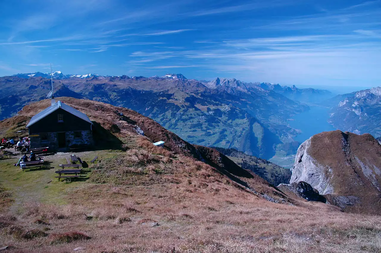 Auf dem Bild sieht man links die Gipfelhütte auf dem Alvier, vor der einige Wanderer an Bänken und Tischen sitzen. Rechts am Bildrand sieht man den Walensee, der ein wenig vom Gipfel des Chli Alvier verdeckt wird. Im Hintergrund sind viele Gipfel der Glarner Alpen zu sehen. Besonders markant sind der vergletscherte Tödi (rechts von der Antenne der Alvierhütte), der Glärnisch in der Bildmitte und etwas weiter rechts der felsig Mürtschenstock. Der Himmel ist blau und mit vielen Schleierwolken verziert. Es ist ein wunderschöner Tag im Oktober.