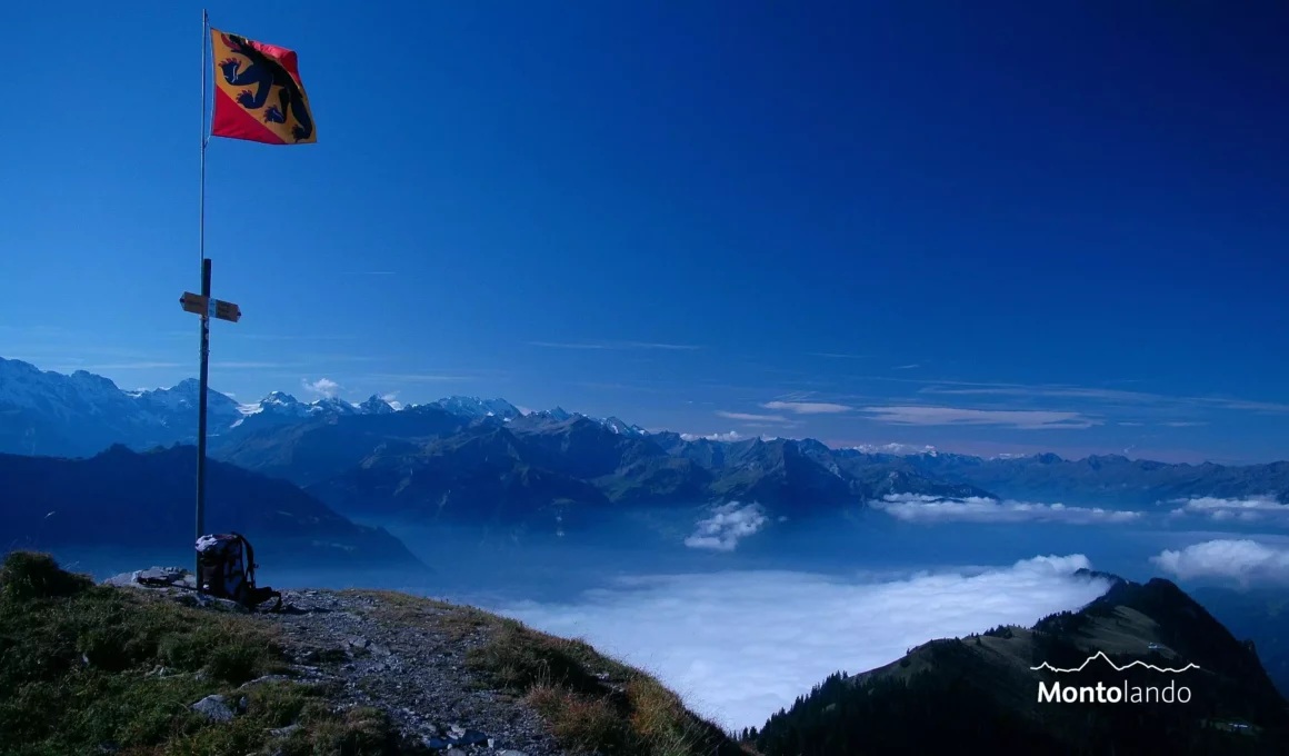 Auf dem Bild sieht man einen hohen Masten mit einer Fahne, auf der das Wappen des Kantons Bern mit einem Bären zu sehen ist. Am Mast ist etwa in der Mitte ein Wegweiser der Schweizer Wanderwege angebracht. Am Boden steht ein grauer Rucksack. Aufnahmestandpunkt ist der Suggiture, ein Vorgipfel des Augstmatthorns. Im Hintergrund sieht man viele Gipfel der Berner Alpen und rechts unter den Grat zum Harder Kulm. Der Brienzersee liegt unter einer Nebelschicht. Der Himmel ist blau, es hat ganz wenige Schleierwolken.