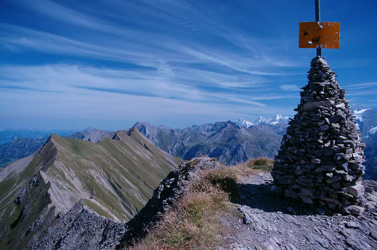 Auf dem Bild geht der Blick vom Bunderspitz Richtung Osten. Rechts im Bild steht ein sehr großer Steinmann, aus dem eine Stange ragt, der die Aussicht auf die Blüemlisalp verdeckt. Links vom Steinmann sind jedoch Jungfrau, Mönch und Eiger zu sehen. Dazu viele Gipfel der Berner Voralpen. Links im Mittelgrund zieht ein grasiger Grat, der Allmegrat zum First über Kandersteg. Der Himmel ist blau und mit wunderschönen Schleierwolken bedeckt.