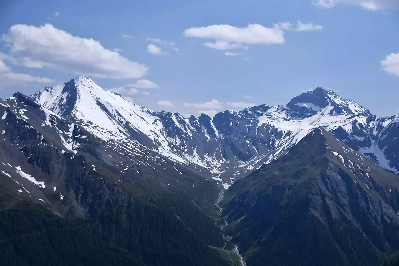 Auf dem Bild geht der Blick vom Piz Munschuns, nördlich oberhalb von Samnaun in Richtung Süden zu den beiden höchsten Bergen der Samnaungruppe, dem Muttler links und dem Stammerspitz rechts. Beide sind Ende Juni noch in weiten Teilen schneebedeckt. Dies gilt vor allem für den Muttler, der in der Gipfelregion fast noch ganz weiß ist. Das liegt daran, dass er aus weichem Schiefer aufgebaut ist und keine steilen Wände hat, wie der Stammerspitz nebenan. Zwischen den beiden Gipfeln ist die Val Maisas mit dem gleichnamigen Bach eingekerbt. Der Himmel ist blau, es hat aber viele kleine Schönwetterwolken und dünne Schleierwolken.