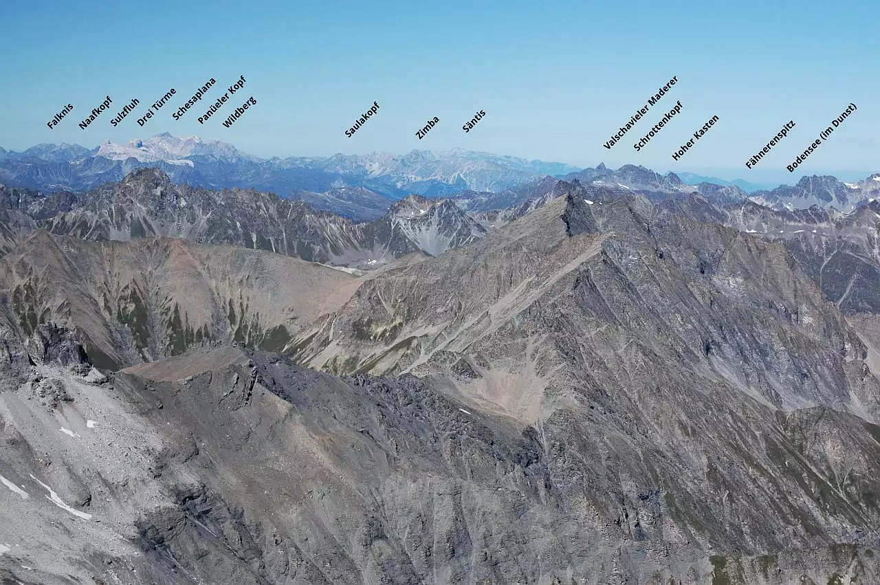 Auf dem Bild geht der Blick vom Muttler über die kahlen Gipfel der Silvretta hinweg zum Rätikon (Sulzfluh, Schesaplana, Zimba), zum Säntis und zum Bodensee, der sich unter einer Dunstschicht verbirgt. Es ist ein außergewöhnlich klarer Tag, selbst der nur 1500 m hohe Fähnerenspitz im Appenzellerland ist bestens zu erkennen. Der Himmel ist blau und wolkenlos.