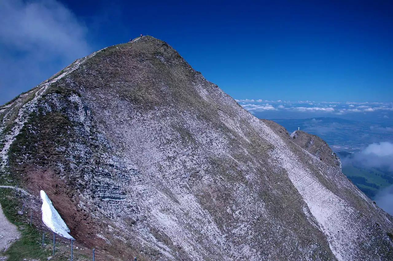 Auf dem Bild geht der Blick von etwas unterhalb der Bergstation zum Gipfel des Moléson. Er bricht nach Osten (rechts) mit sehr steilen Grasschrofen ab. Am linken Bildrand sieht man den nur wenig steilen und breiten Südgrat, über den der Weg zum Gipfel führt. Die Wegtrasse ist sehr gut zu sehen, ebenso einige Wanderer, die zum Gipfel unterwegs sind. Im Sattel unterhalb des Gipfels liegt noch ein kleines Altschneefeld. Rechts sieht man im Hintergrund noch ein Kreuz auf einem Vorgipfel und dahinter das Mittelland in der Umgebung von Bulle. Dort sind ebenso einige Nebelfelder zu sehen, wie links vom Gipfel. Abgesehen von wenigen Wolken ist der Himmel blau.