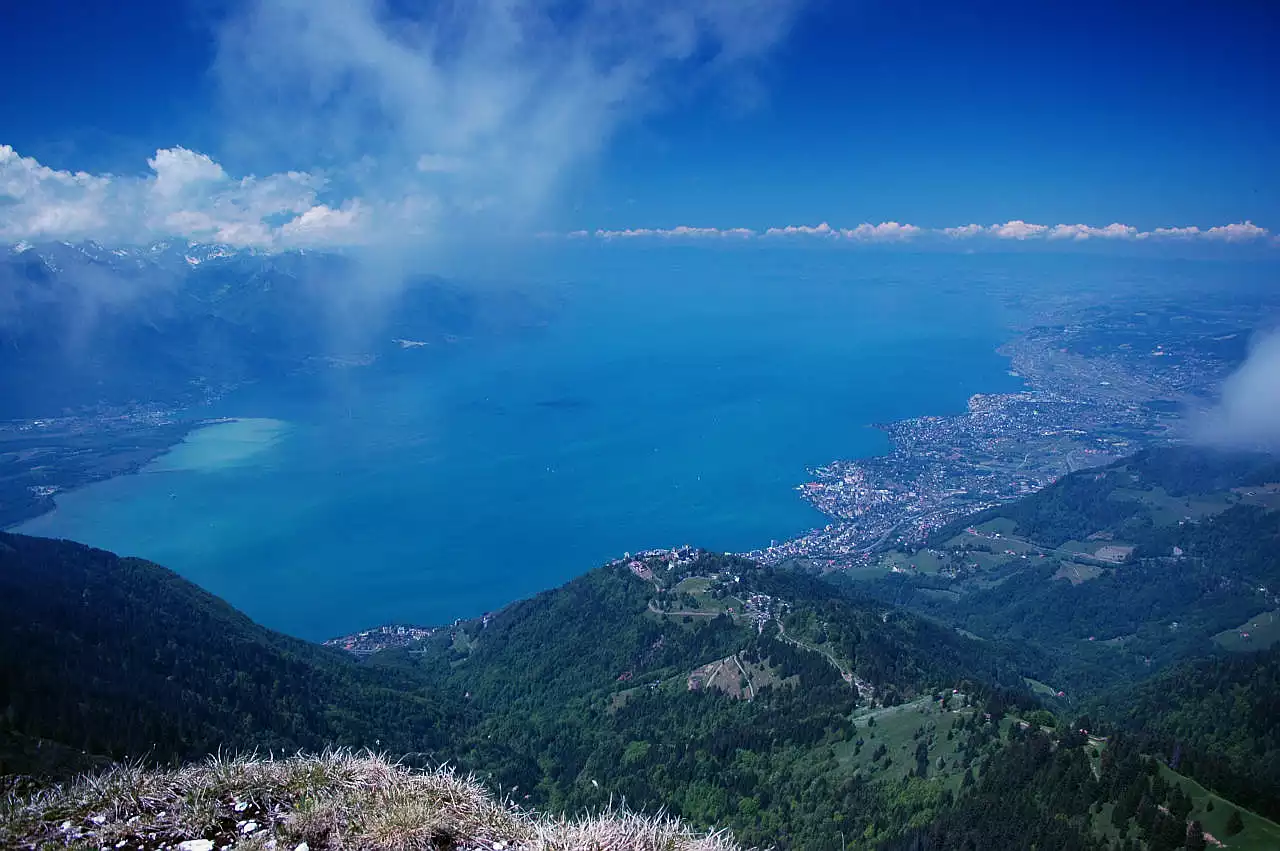 Genfersee. Er ist nicht in seiner ganzen Ausdehnung sichtbar. Rechts am Ufer erkennt man die Städte Vevey und Montreux, auf einem Hügel unterhalb sieht man die Ortschaft Caux. Links über dem See erhebt sich die Berge zwischen Le Grammont und Dent d'Oche, die größtenteils in Wolken stecken. Auch rechts sieht man am Horizont über dem Jura große Quellwolken. Darüber ist der Himmel blau.