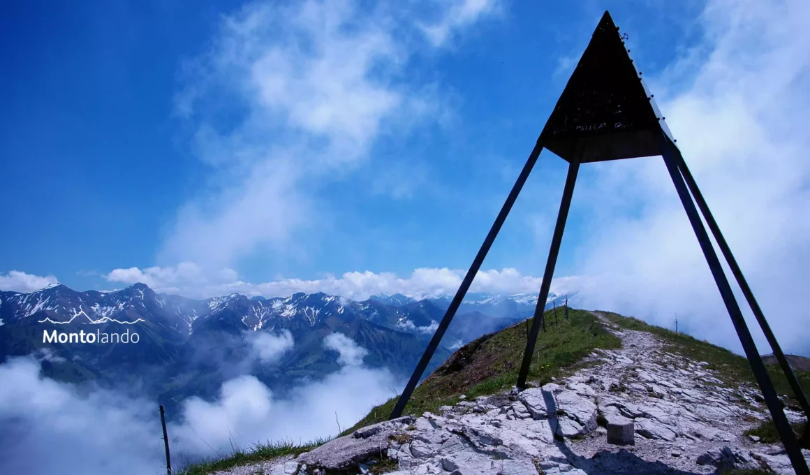 Auf dem Bild sieht man das trigonometrische Signal in Form einer Pyramide am Moléson. Dahinter erkennt man den letzten Abschnitt des Wegs, der von der Bergstation auf den Gipfel führt. Links sieht man im Hintergrund den südlichsten Abschnitt der höchsten Kette der Freiburger Voralpen mit den Gipfel Vanil de l'Ecri und Pointe de Paray. Beide Berge sind zum Teil noch mit Schneefeldern bedeckt. Die Gipfel der Waadtländer Alpen am Horizont stecken hingegen in Wolken. Ebenso liegen in den Tälern Wolken und auch rechts über dem Signal hat es dichte Wolken. Über den Wolken ist der Himmel blau.