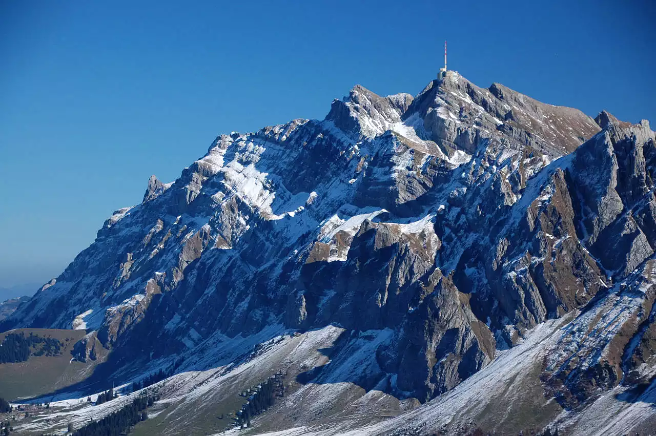 Auf dem Bild geht der Blick vom Stockberg in nordöstlicher Richtung zum Säntis. Der Gipfel erhebt sich unmittelbar über den grünen Wiesen der Schwägalp, die sich am linken Bildrand erstreckt. Die Säntis ist besonders auf der Nordseite (links) verschneit und auch im Vordergrund sieht man ein paar Neuschneeflecken. Der Himmel ist blau und wolkenlos an einem Traumtag Ende Oktober.