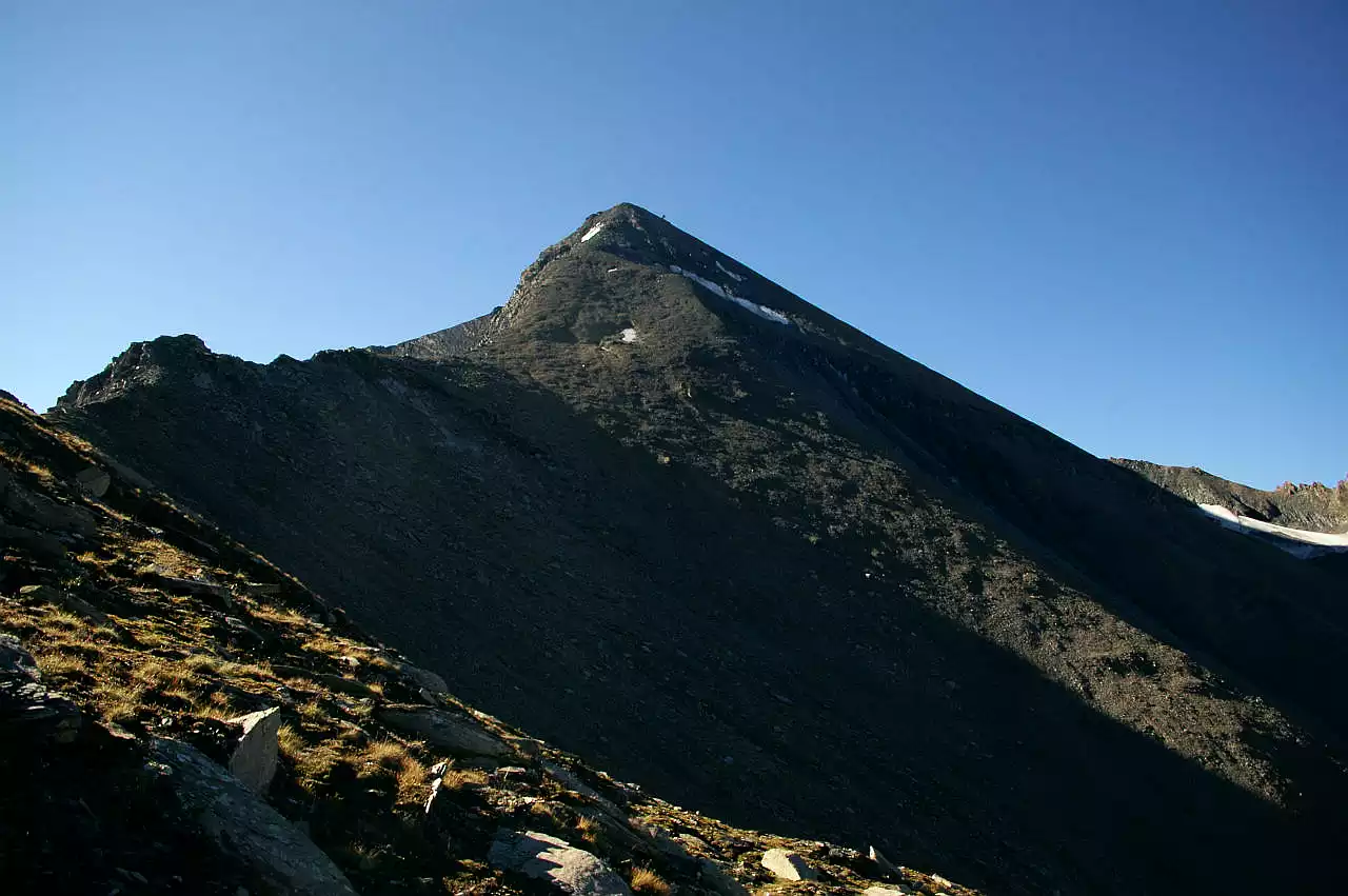 Auf dem Bild sieht man den Gipfelkopf des Muttlers von Norden. Nach rechts fällt die Nordwestflanke, die zum Teil noch im Schatten liegt, steil in die Val Maisas ab. Nach links zieht der Nordgrat zum Gipfel, der nach oben hin immer steiler wird. Ein paar kleinere Schneefelder halten sich noch rund um den Gipfel. Der Himmel ist blau und wolkenlos.