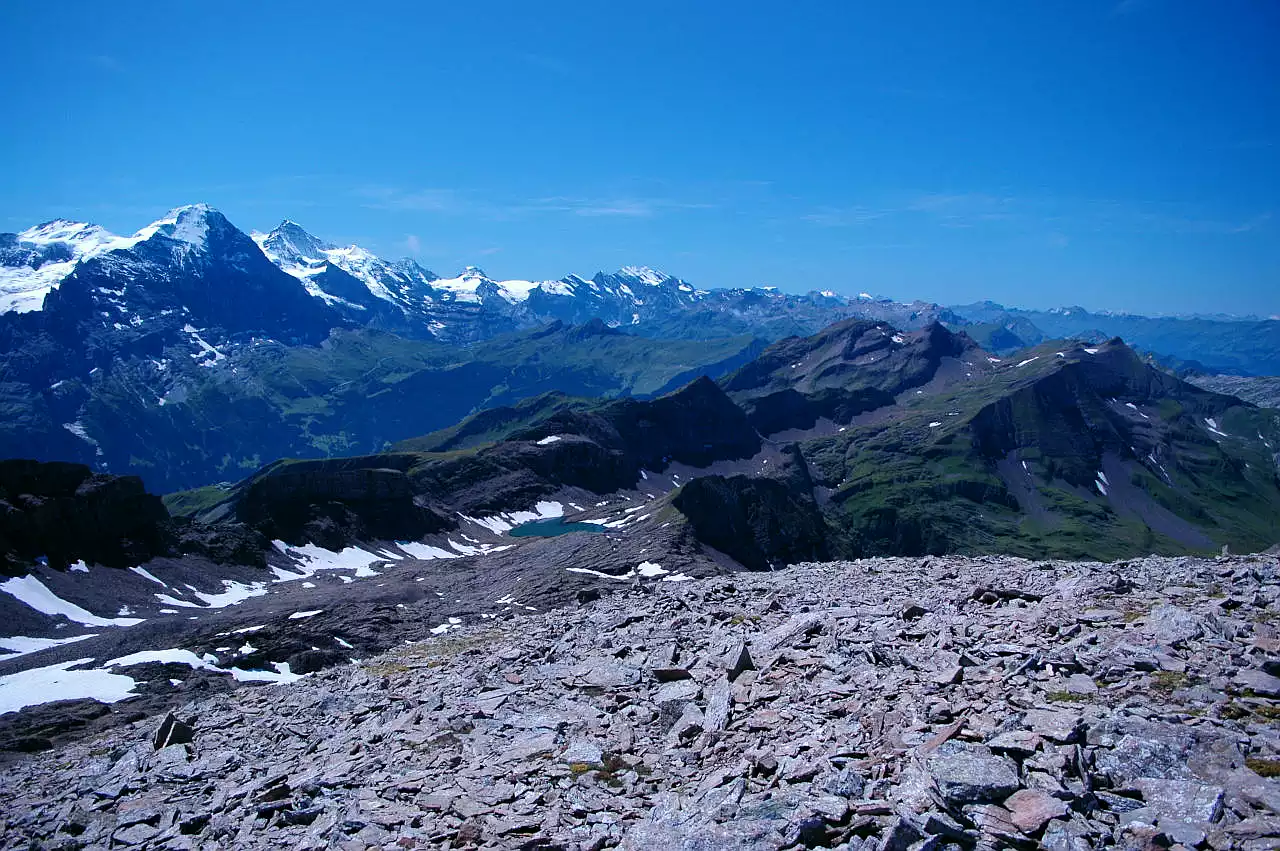 Auf dem Bild geht der Blick vom Wildgärst Richtung Südwesten zu den Gipfeln der Berner Alpen. Links sieht man die vergletscherten Hochalpen vom Eiger bis zum Wildhorn, das nur noch ganz schwach am Horizont zu sehen ist. Nach rechts schließen sich die niedrigeren, teils begrünten Berner Voralpen an. Ziemlich weit rechts im Bildmittelgrund ragt daraus die kleine Spitze des Faulhorns heraus. Im Vordergrund liegen viele Kalksteine, die den Gipfel des Wildgärst bedecken. Der Himmel ist blau, es hat ganz wenige Schleierwolken.