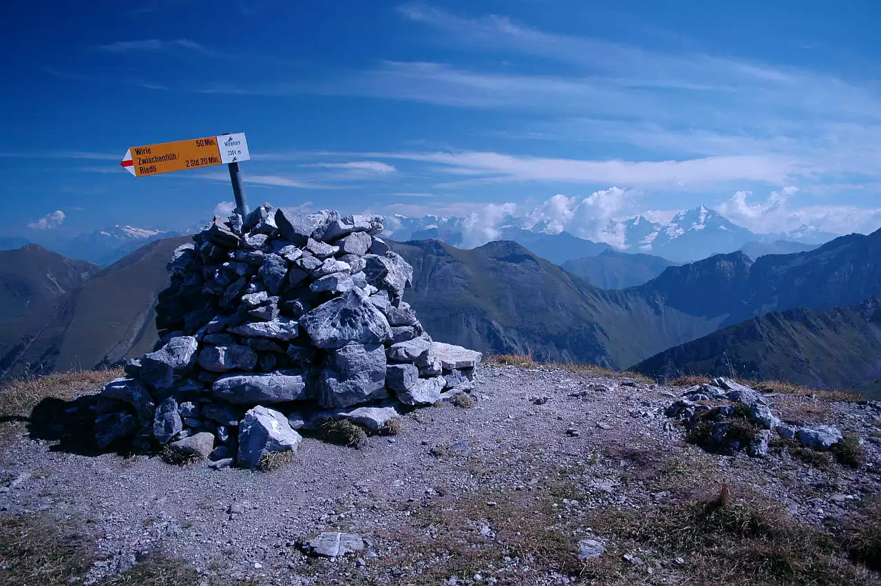 Auf dem Bild sieht man den breiten Gipfelsteinmann auf dem Wiriehorn. Darin steckt ein Wegweiser der Schweizer Wanderwege, auf dem auch die Ortsangabe "Wiriehorn" zu lesen ist. Im Hintergrund sieht man die grasigen Gipfel der Niesenkette, überragt von den Berner Hochalpen zwischen Wetterhorn, links und Doldenhorn, rechts. Viele der Gipfel stecken in Quellwolken. Darüber ist der Himmel blau, mit wenigen Schleierwolken.