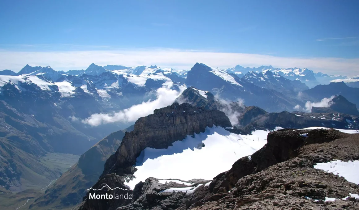 Auf dem Bild geht der Blick vom Wissigstock Richtung Südwesten zu den Gipfeln der Urner Alpen, mit Fleckistock, Sustenhorn, Dammstock und Gwächtenhorn links und in der Bildmitte sowie den Berner Alpen, mit Finsteraarhorn, Schreckhorn und Wetterhörnern rechts. Knapp rechts der Bildmitte fällt der Titlis mit seiner Gletscherkappe auf. Darunter sieht man den Gipfel des Schuflen im Vordergrund. Zu sehen ist rechts davon die Bahnstation am Chli Titlis. Links unten erkennt man in der Tiefe das Tal von Surenen. Der Himmel ist blau, am Horizont hat es Schleierwolken und um die Gipfel ziehen einzelne Nebelfetzen.