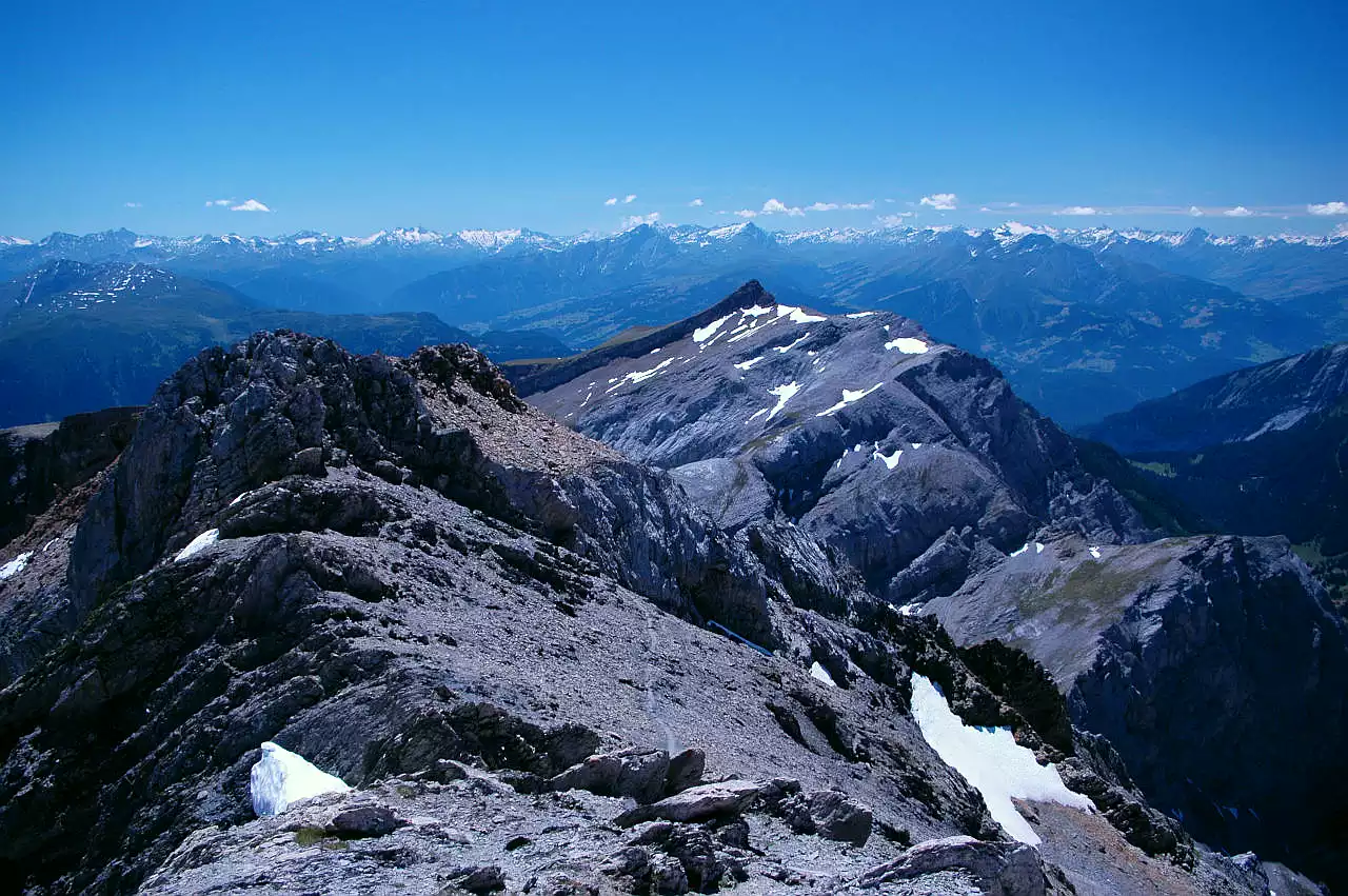 Auf dem Bild geht der Blick vom Gipfel des Haldensteiner Calanda Richtung Süden auf die Bündner Bergwelt mit vielen Gipfeln zwischen Plattgruppe links und Adula rechts. Auf den Nordseiten der Berge liege noch einige Altschneefelder. Im Bildmittelgrund sieht man die Spitze des Felsberger Calanda. Dahinter kann man in der Tiefe das Rheintals erahnen. Im Vordergrund erkennt man im Schutt die Wegspur, die von der Calandahütte zum Gipfel führt. Der Himmel ist blau mit wenigen Schönwetterwolken.