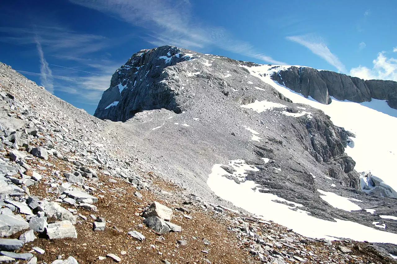 Auf dem Bild sieht man den Gipfelaufbau mit der letzten Etappe zum Wissigstock. Man erkennt der Steig, der durch den Kalkschutt auf er westlichen Seite des Grates zum Gipfel führt. Auf dieser Seite sind die Hänge flacher, während der Berg Richtung Osten (links) mit steilen Wänden abbricht. Am rechten Bildrand ist in einer Mulde noch des Griessenfirns zu sehen. Der Himmel ist blau, es hat wenige Schleierwolken.